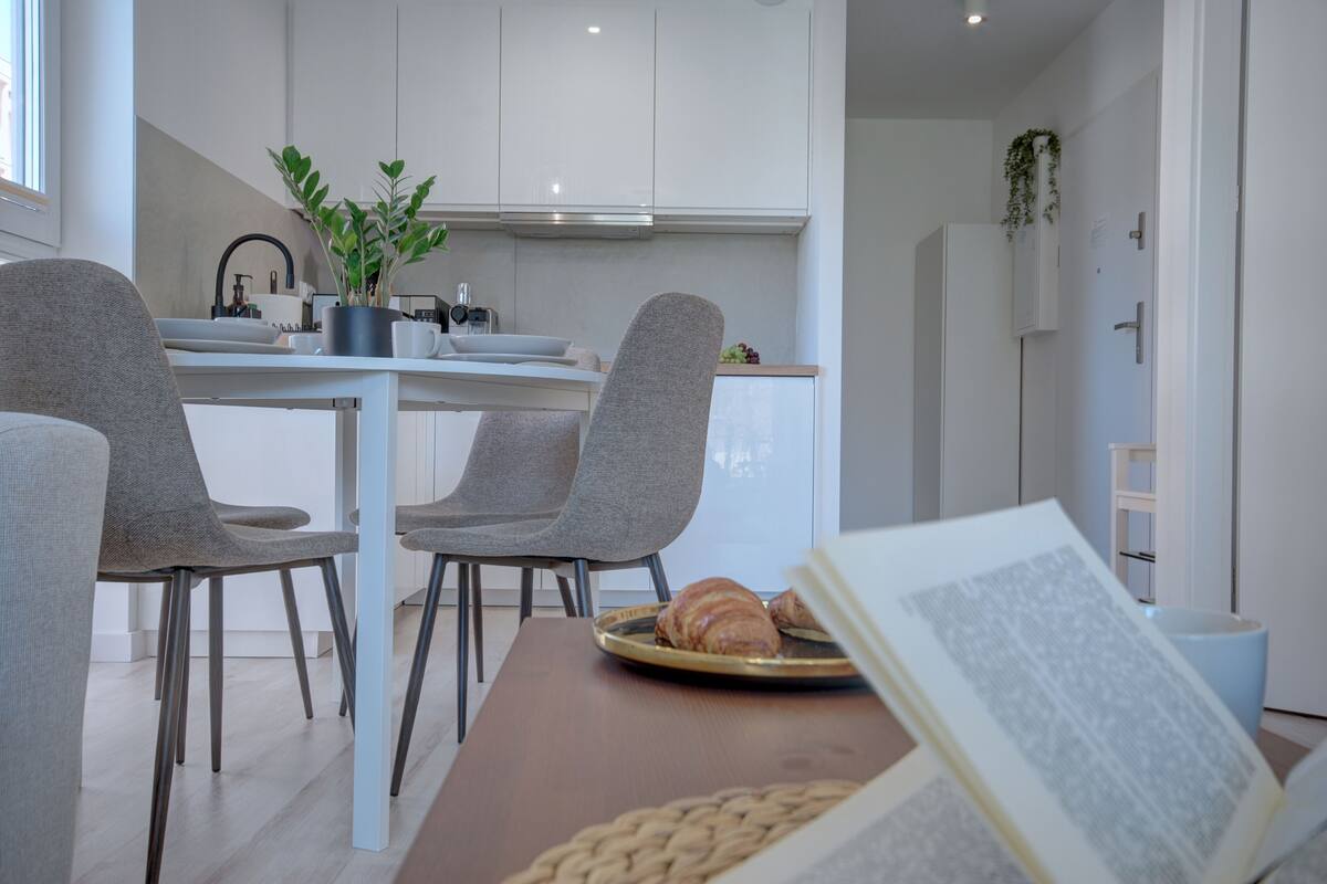 Una cucina moderna con mobili bianchi, un'area pranzo con sedie grigie e un libro aperto su un tavolo.