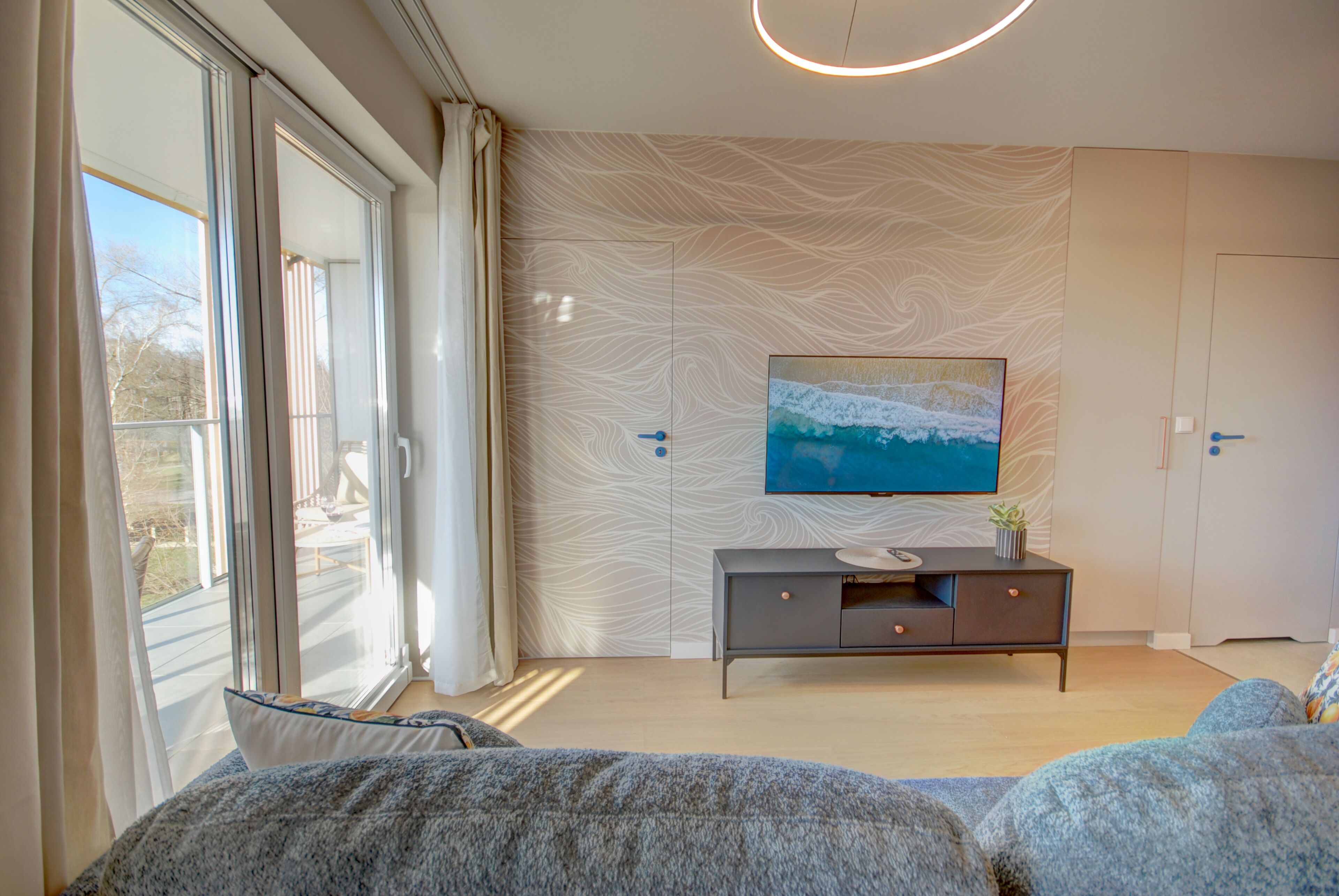 A modern living room with a large window, a flat-screen TV on the wall, a grey sofa, and a wooden floor.