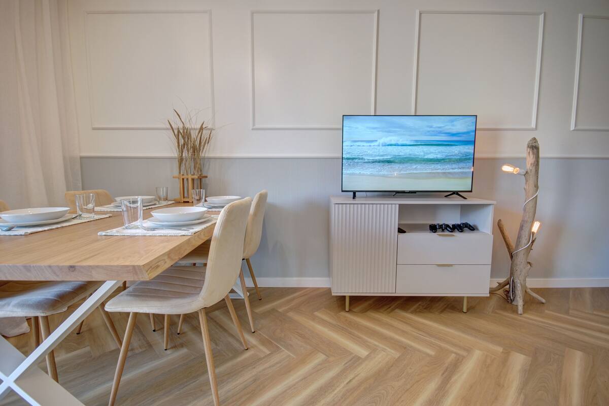 Zona pranzo con tavolo in legno, sedie bianche e televisore a schermo piatto su un mobile bianco.