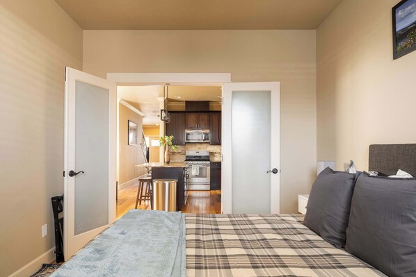 A bedroom with a bed, a bench, and a view of a kitchen and dining area.