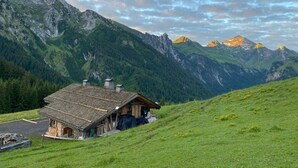Una cabaña rústica de montaña con techo inclinado, rodeada de exuberantes campos verdes y montañas imponentes bajo un cielo parcialmente nublado.