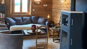 Interior acogedor de una cabaña de troncos con un sofá, una silla de madera, una mesa con una taza, una chimenea y una ventana con vistas al exterior.