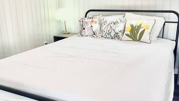 Una cama bien hecha con colchón blanco y estructura de metal negro, una mesita de noche con lámpara y una ventana con vistas a árboles.