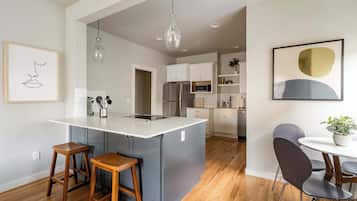 Een moderne keuken met een centraal eiland, houten krukken, een eettafel met stoelen en een hangende lamp.