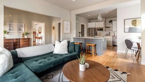 A modern living room with a dark green sectional sofa, a round wooden coffee table, and a kitchen in the background.