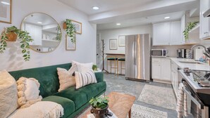 A modern living room with a green sofa, a coffee table, and a kitchen area with white cabinets and stainless steel appliances. - Retro Hideaway - Trendy Location - 2BR - Sleeps 4 (Portland)