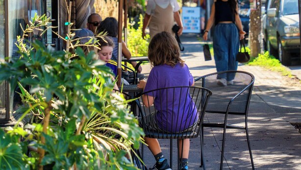Un caffè all'aperto con tavoli e sedie sotto un ombrello verde, una strada con passanti e auto parcheggiate.
