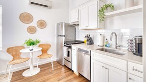 Una cucina moderna con mobili bianchi, un frigorifero in acciaio inossidabile, un fornello e una lavastoviglie. C'è anche un piccolo angolo ristoro con sedie in vimini e un tavolo.