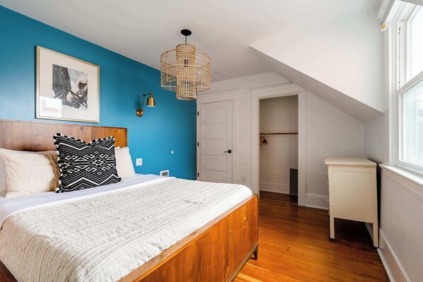 A bedroom with a wooden bed, a white bedside table, a blue accent wall, a hanging light fixture, and a framed picture on the wall.