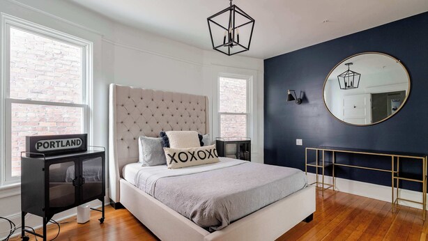 A bedroom with a large bed, a bedside table, a dresser, a mirror, and a window with a view of a brick building.