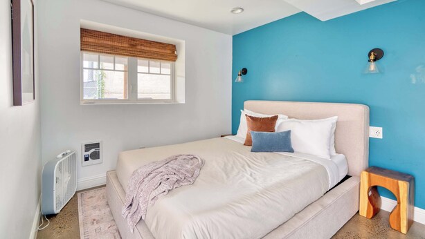 A bedroom with a bed, a window with blinds, a standing fan, and a blue accent wall. - Massive 7BR - Nob Hill - NW 23rd - Sleeps 14 (Portland)