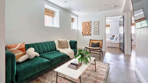 A spacious living room with a green sofa, a coffee table, and a plant. There are two windows with blinds, a wall-mounted artwork, and a door leading to another room. - Massive 7BR - Nob Hill - NW 23rd - Sleeps 14 (Portland)