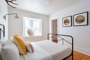 Un dormitorio con cama, ventana, lámpara de pared y dos cuadros enmarcados.