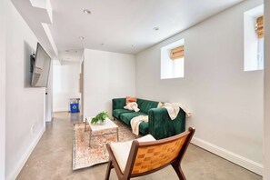 Uma sala de estar moderna com um sofá verde, uma cadeira de madeira, uma mesa pequena com uma planta e um tapete decorativo.