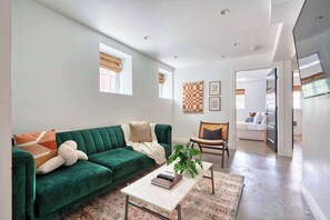 Ein geräumiges Wohnzimmer mit einem grünen Sofa, einem Couchtisch und einer Pflanze. Zwei Fenster mit Jalousien, ein Wandbild und eine Tür zu einem anderen Raum.