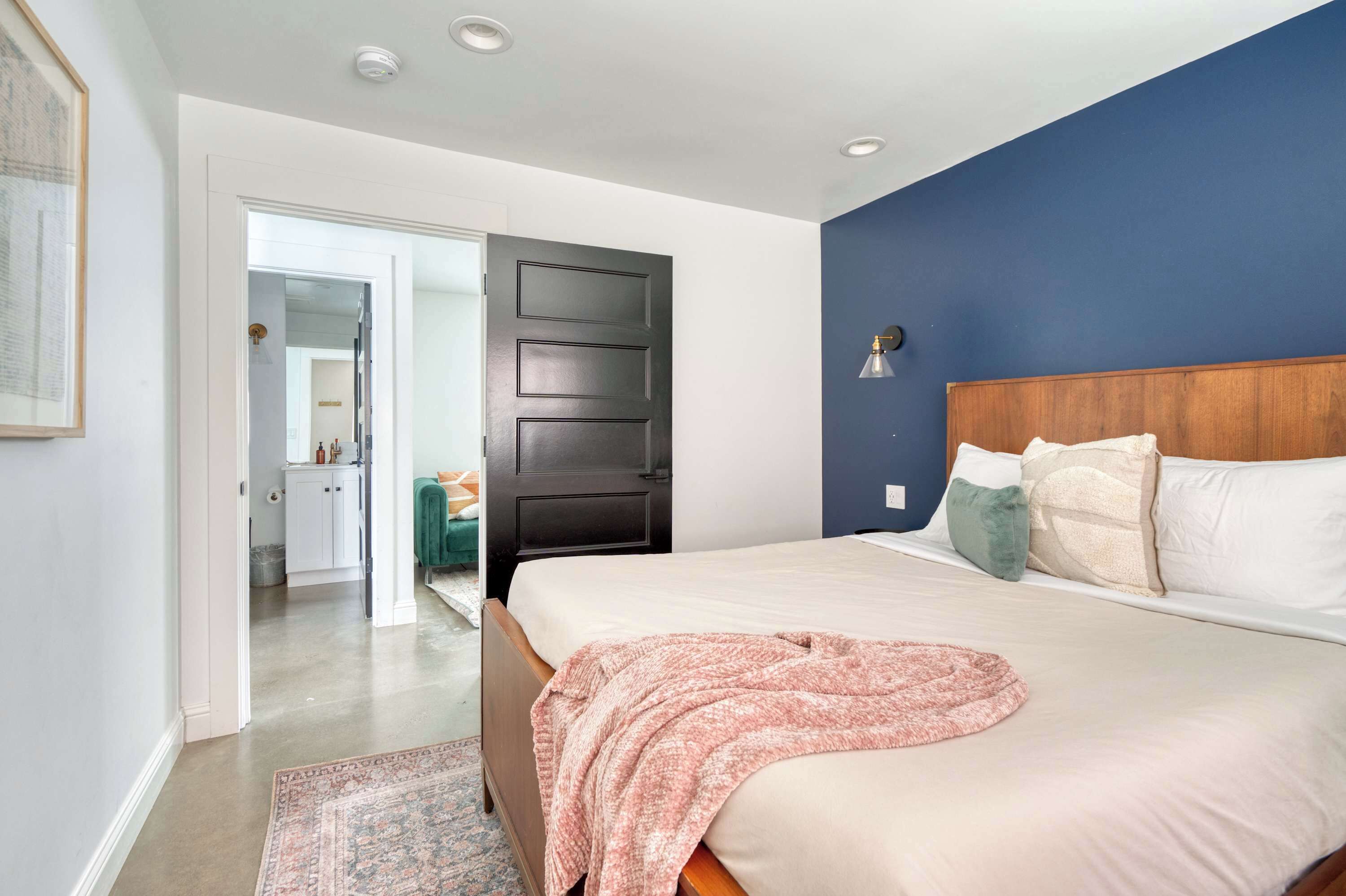 A bedroom with a large bed, a dark door, a blue accent wall, and a hallway leading to a bathroom.