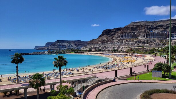 Una playa costera con un paseo marítimo curvo, palmeras y montañas al fondo.