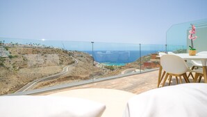 A terrace with a view of a coastal road and ocean. There is a table with chairs and a potted plant.