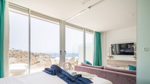 A modern hotel room with a large bed, a sofa, a TV, and a balcony with a view of the coastline.