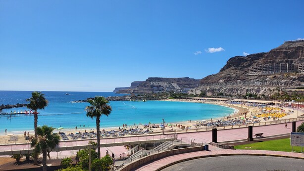 A coastal resort with a large sandy beach, a promenade, and a rocky cliff backdrop.