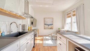 A modern kitchen with white cabinetry, a stainless steel sink, and a wooden countertop. There is a window with curtains and a painting on the wall. - 1BR • Fast WiFi • Walk to Dining & Breweries (Portland)