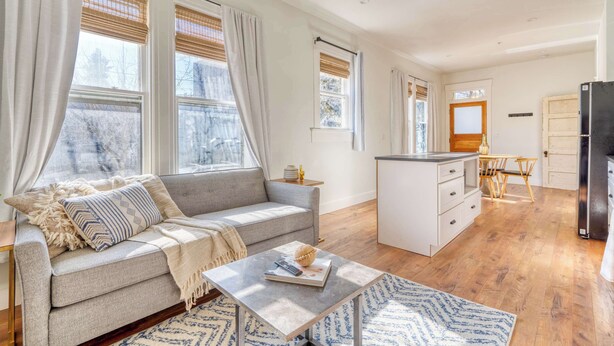 A modern apartment with a grey sofa, a small coffee table, and a kitchen area with wooden flooring. - 1BR • Fast WiFi • Walk to Dining & Breweries (Portland)