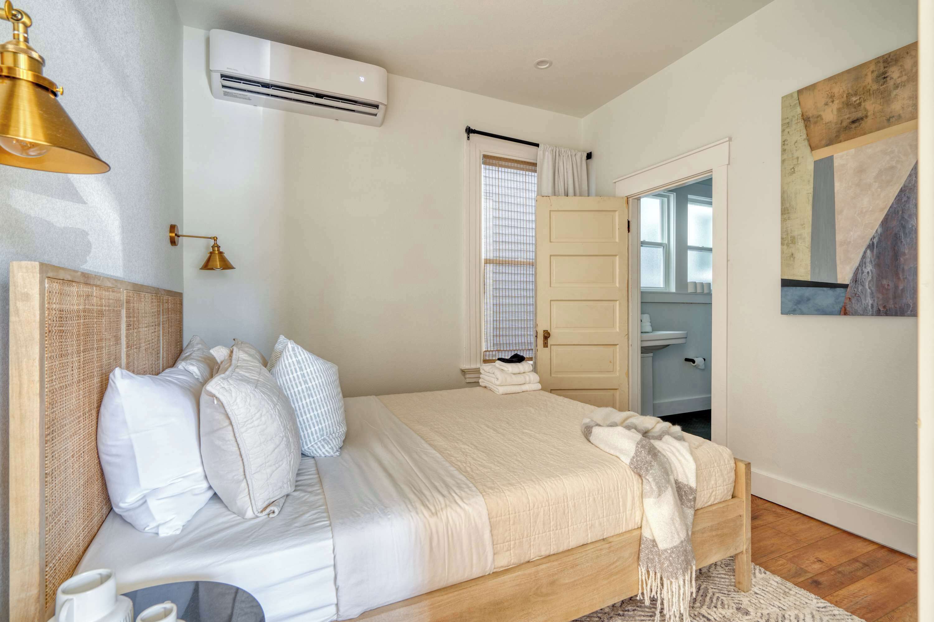 A bedroom with a bed, bedside lamp, air conditioning unit, window with blinds, and a door leading to a bathroom.