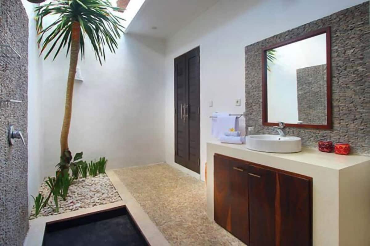Een moderne badkamer met een witte wastafel, donkere houten kastjes, een stenen wand en een kleine palmboom.