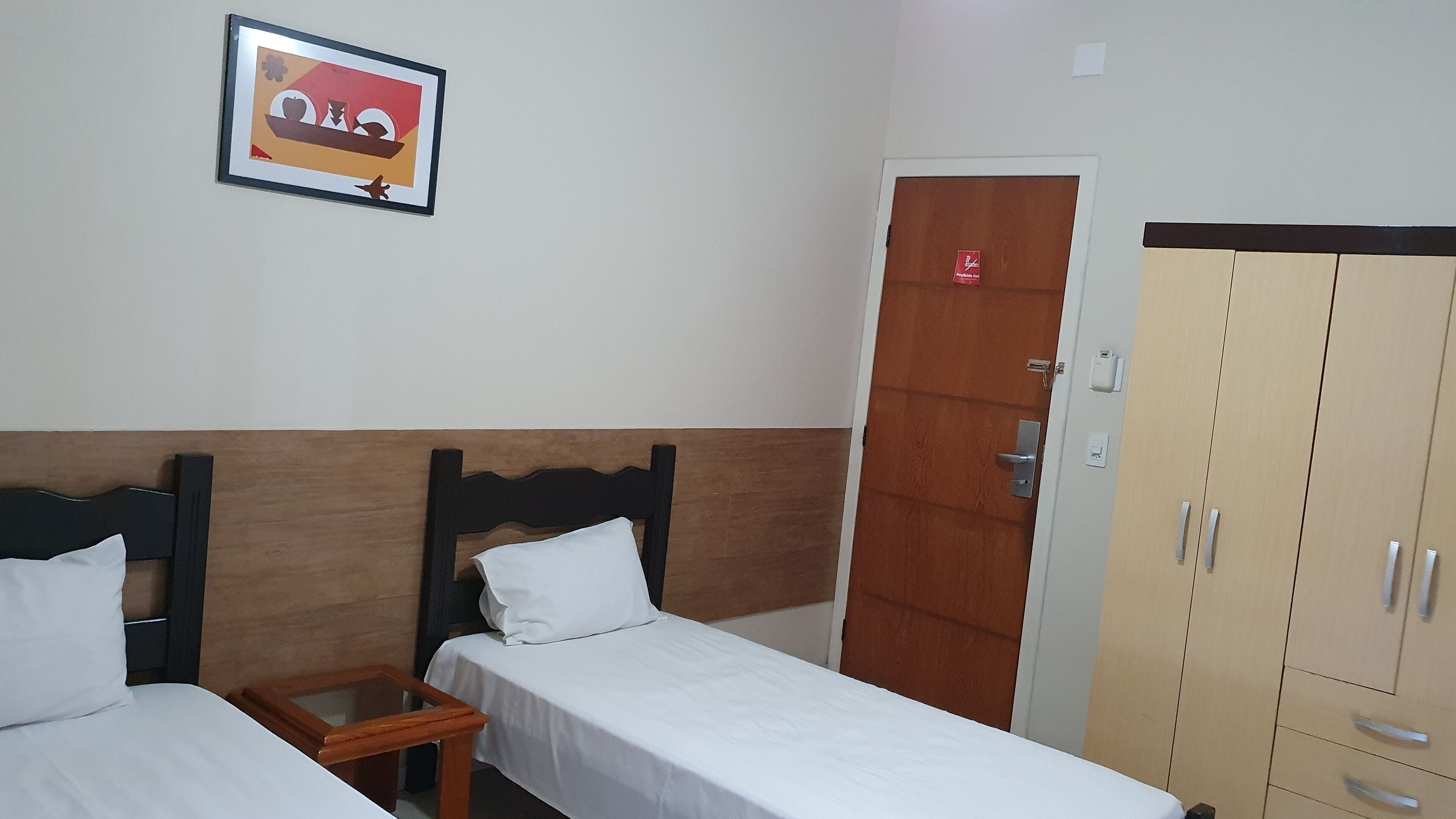 Habitación de hotel con dos camas individuales, cabecero de madera, mesita de noche de madera, un cuadro enmarcado en la pared, armario con varios cajones y una puerta con un letrero rojo.