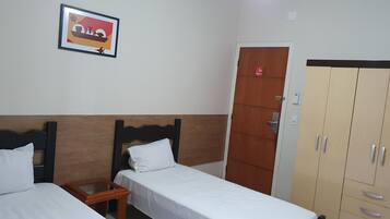 Habitación de hotel con dos camas individuales, cabecero de madera, mesita de noche de madera, un cuadro enmarcado en la pared, armario con varios cajones y una puerta con un letrero rojo.