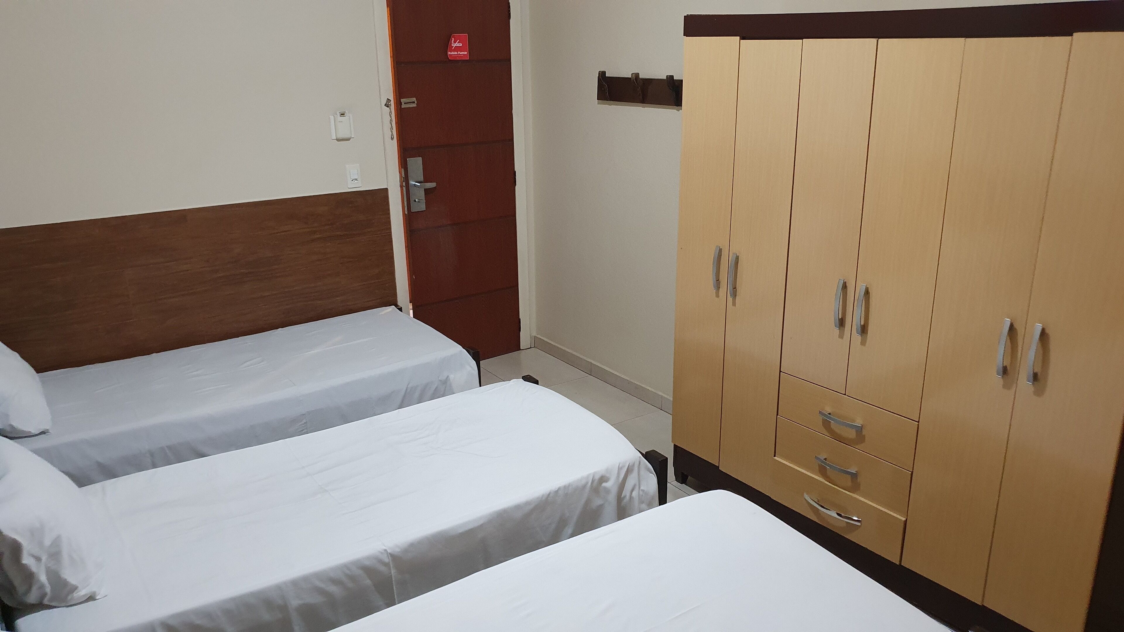 A hotel room with two single beds, a wooden wardrobe, and a door with a red sign.