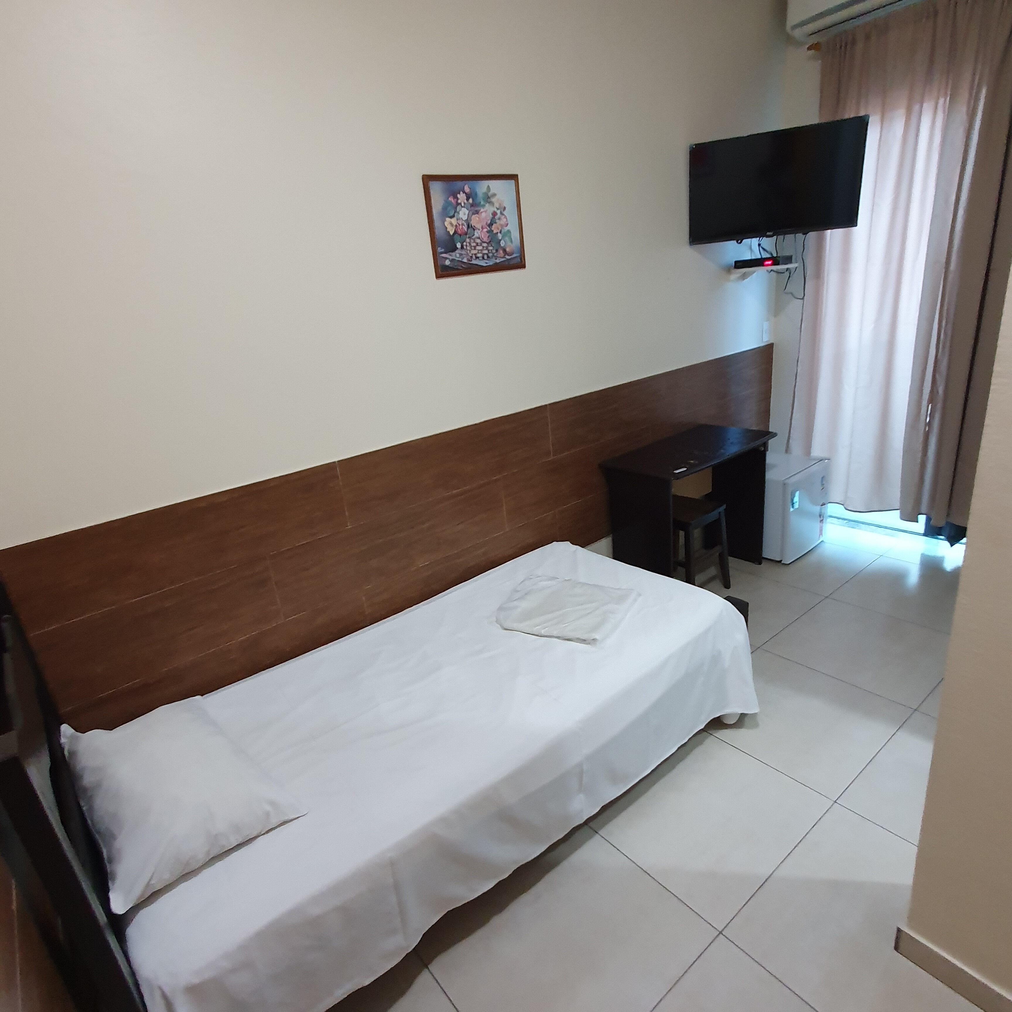A hotel room with a single bed, a desk, a chair, a TV mounted on the wall, and a window with curtains.