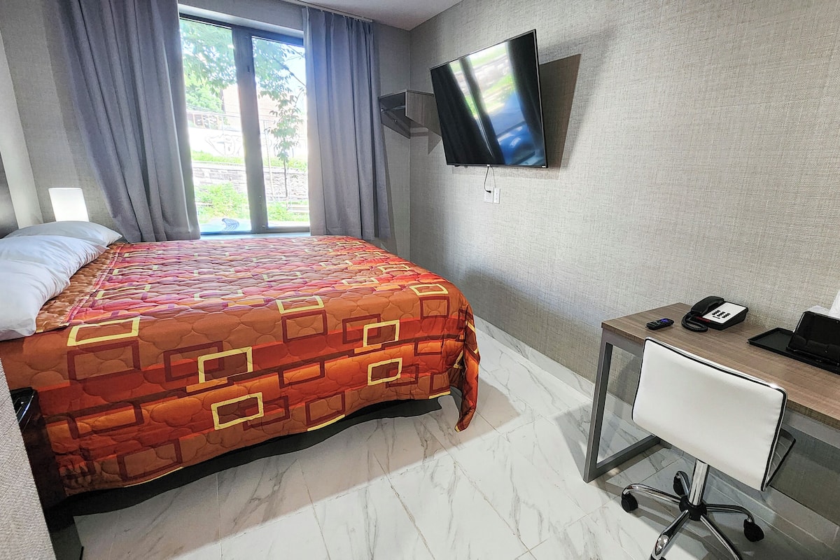 A hotel room with a bed, a desk, a chair, a flat-screen TV mounted on the wall, and a window with curtains.