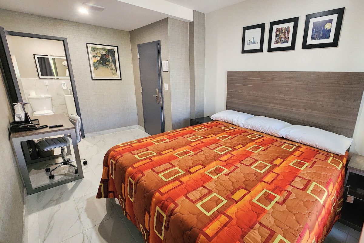 A hotel room with a bed, a desk with a chair, a mirror, and a door. There are three framed pictures on the wall.