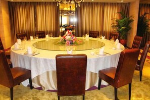 Une table de salle à manger ronde, recouverte d’une nappe blanche, entourée de chaises, dans une pièce agrémentée de rideaux et d’un lustre.