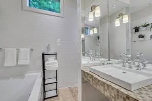 Une salle de bain moderne avec des murs recouverts de carreaux blancs, une baignoire, un meuble-lavabo avec deux vasques et un grand miroir.