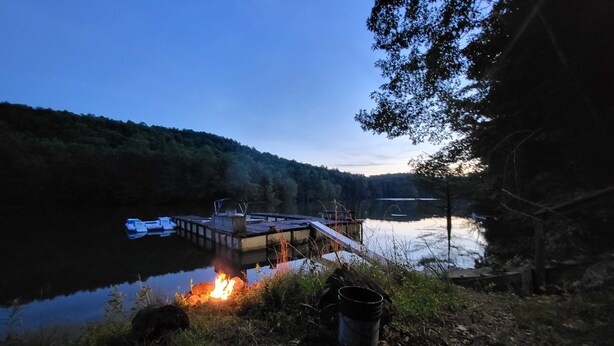 Un muelle que se adentra en un lago tranquilo rodeado de un bosque denso, con una fogata y equipo de campamento en la orilla.