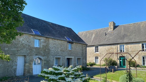 A stone building with multiple windows and a green door, surrounded by a grassy area and a metal bench. - Domaine des Piéris  (Grandcamp-Maisy)