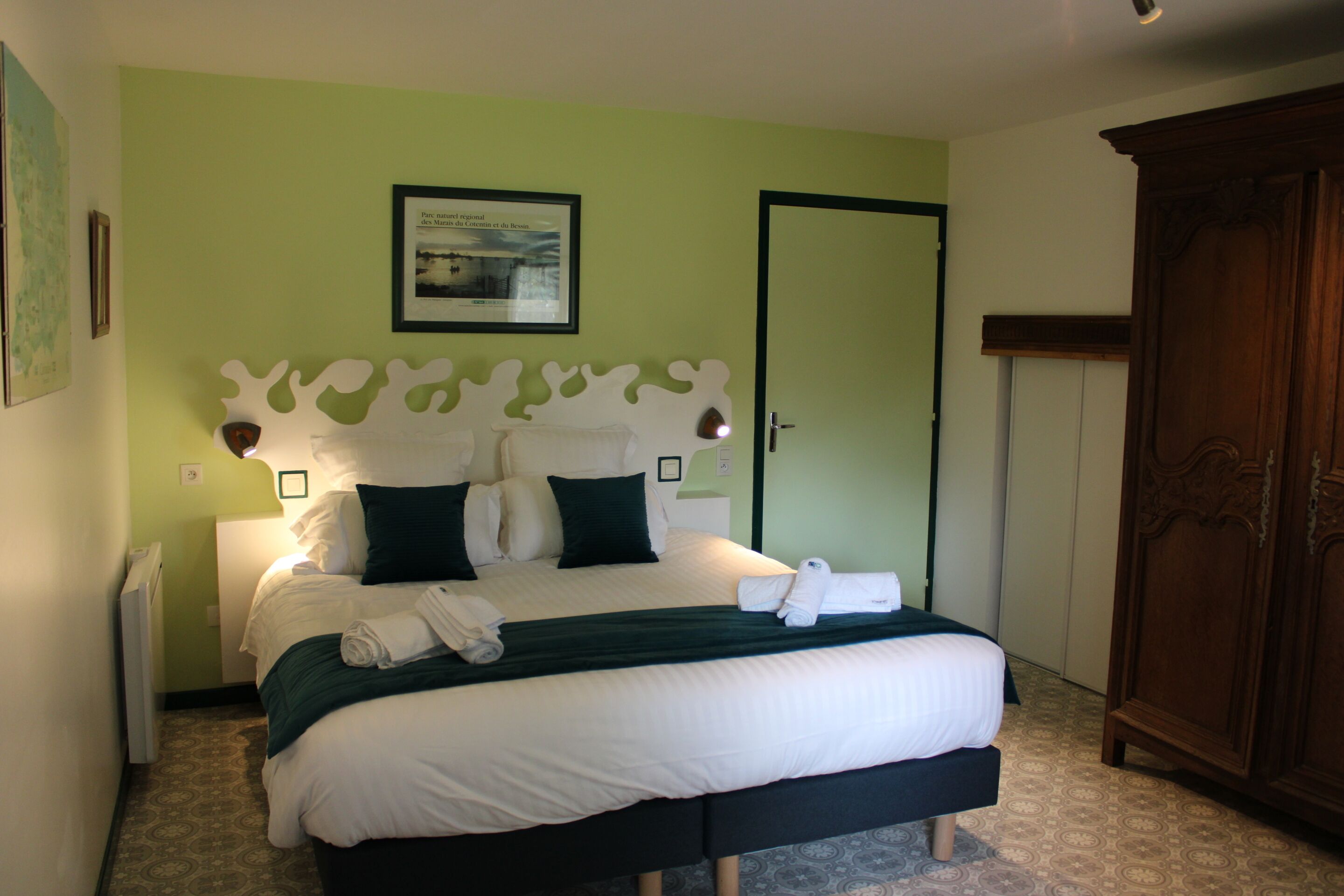 A neatly made bed with white and dark green bedding, a framed picture above it, and a wooden wardrobe to the right.