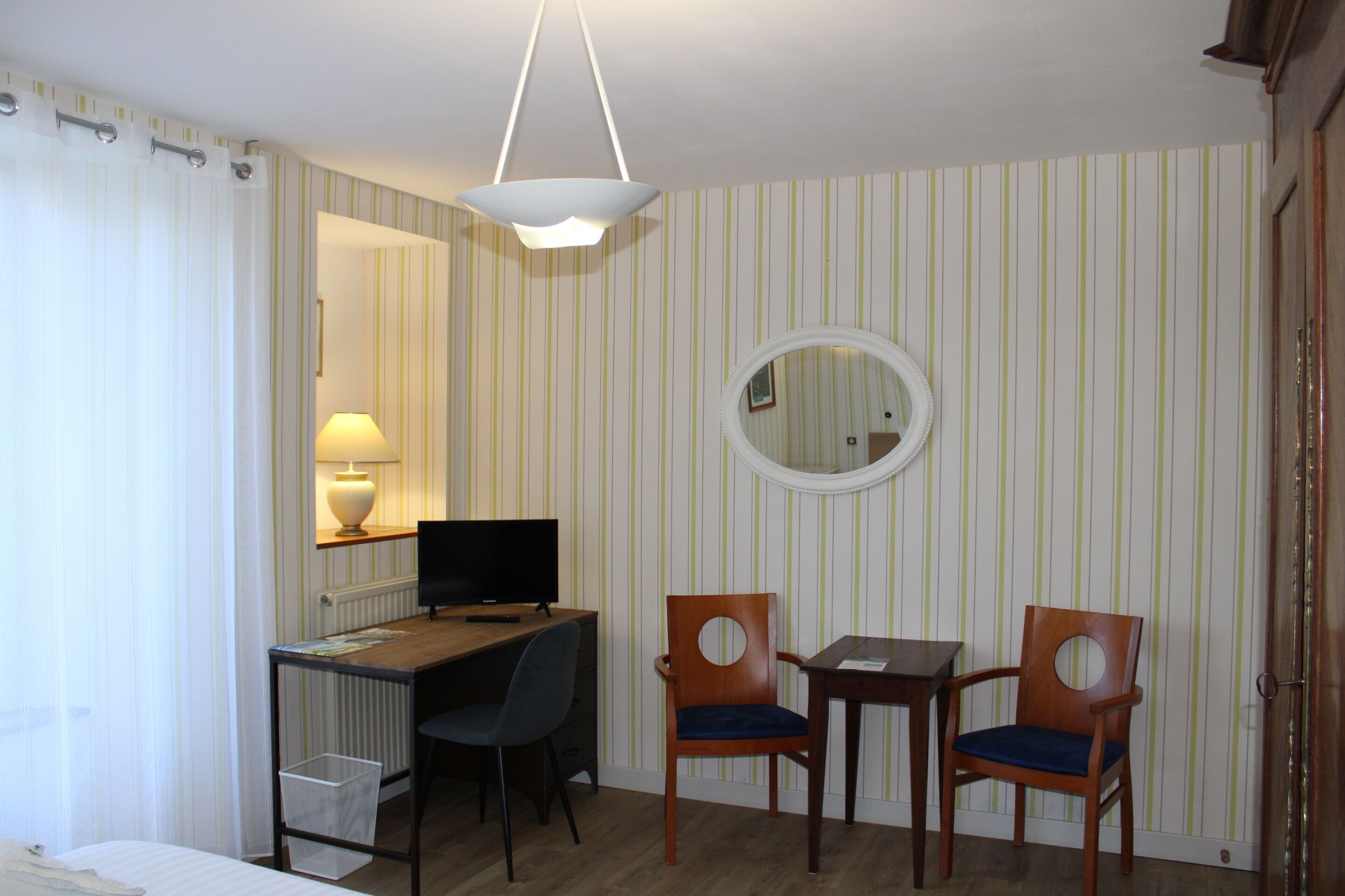 A hotel room with a bed, a desk with a computer, two chairs, a lamp, a round mirror, and striped wallpaper.