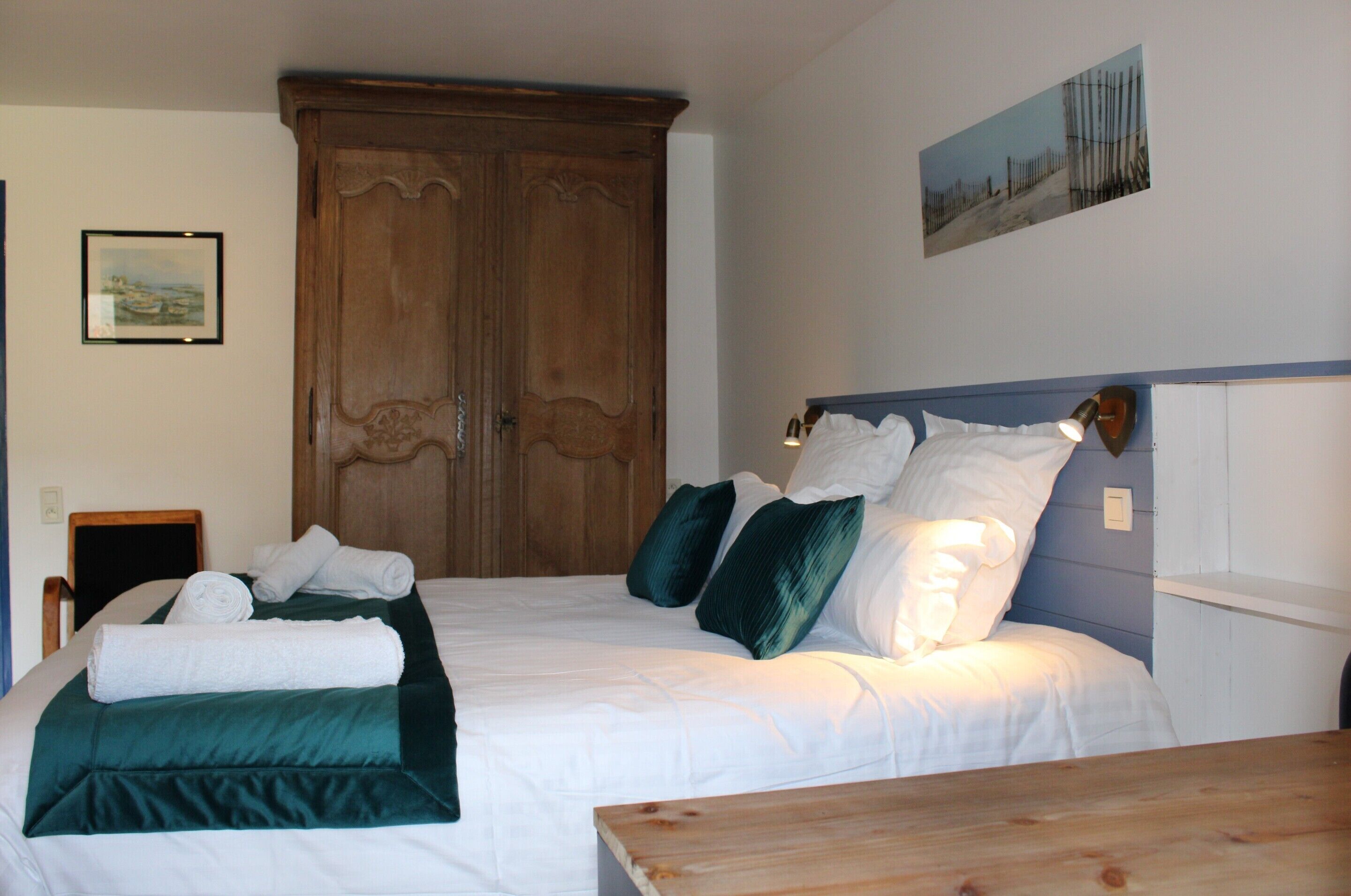 Een netjes opgemaakt bed met witte lakens en groene accenten, een houten garderobe en een lijst met een afbeelding aan de muur.