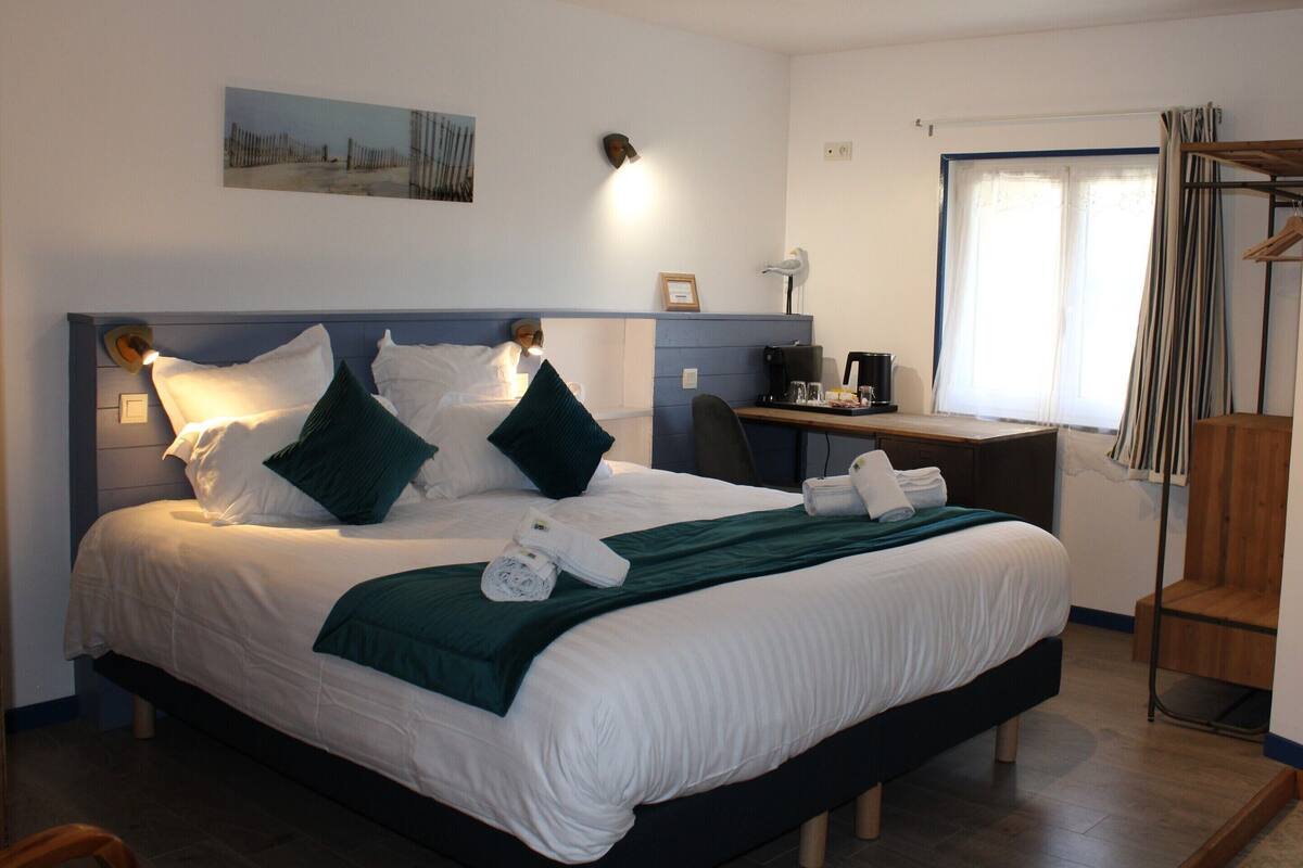 A neatly made bed with white and green bedding, a desk with a computer, and a window with curtains.