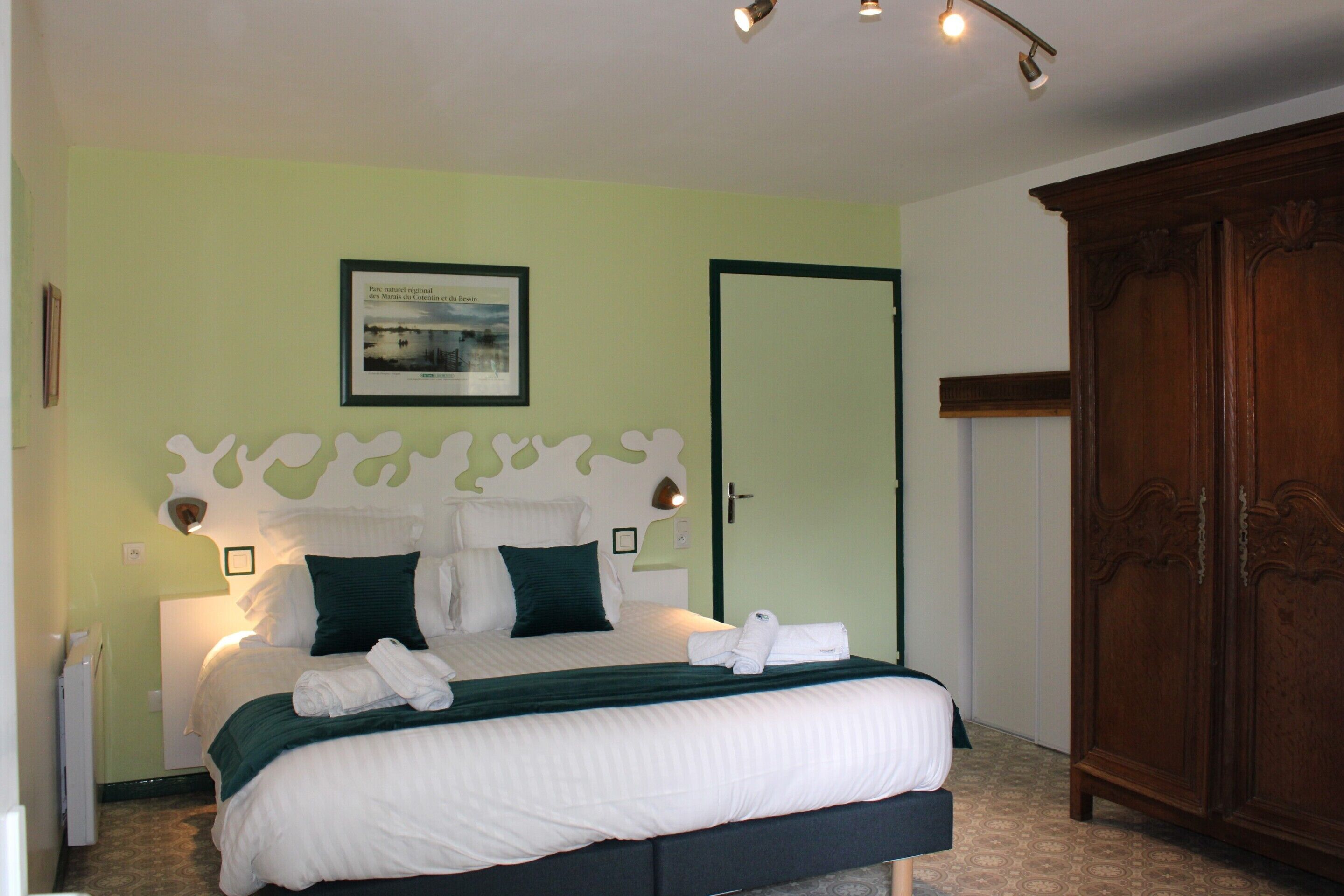 Un lit bien fait, avec du linge de lit blanc et un plaid vert, une armoire en bois et un tableau encadré au mur.