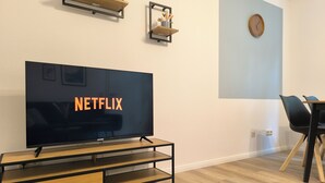 A modern living room with a flat-screen TV displaying 'NETFLIX', a wooden coffee table, a dining area with a table and chairs, and a wall clock.