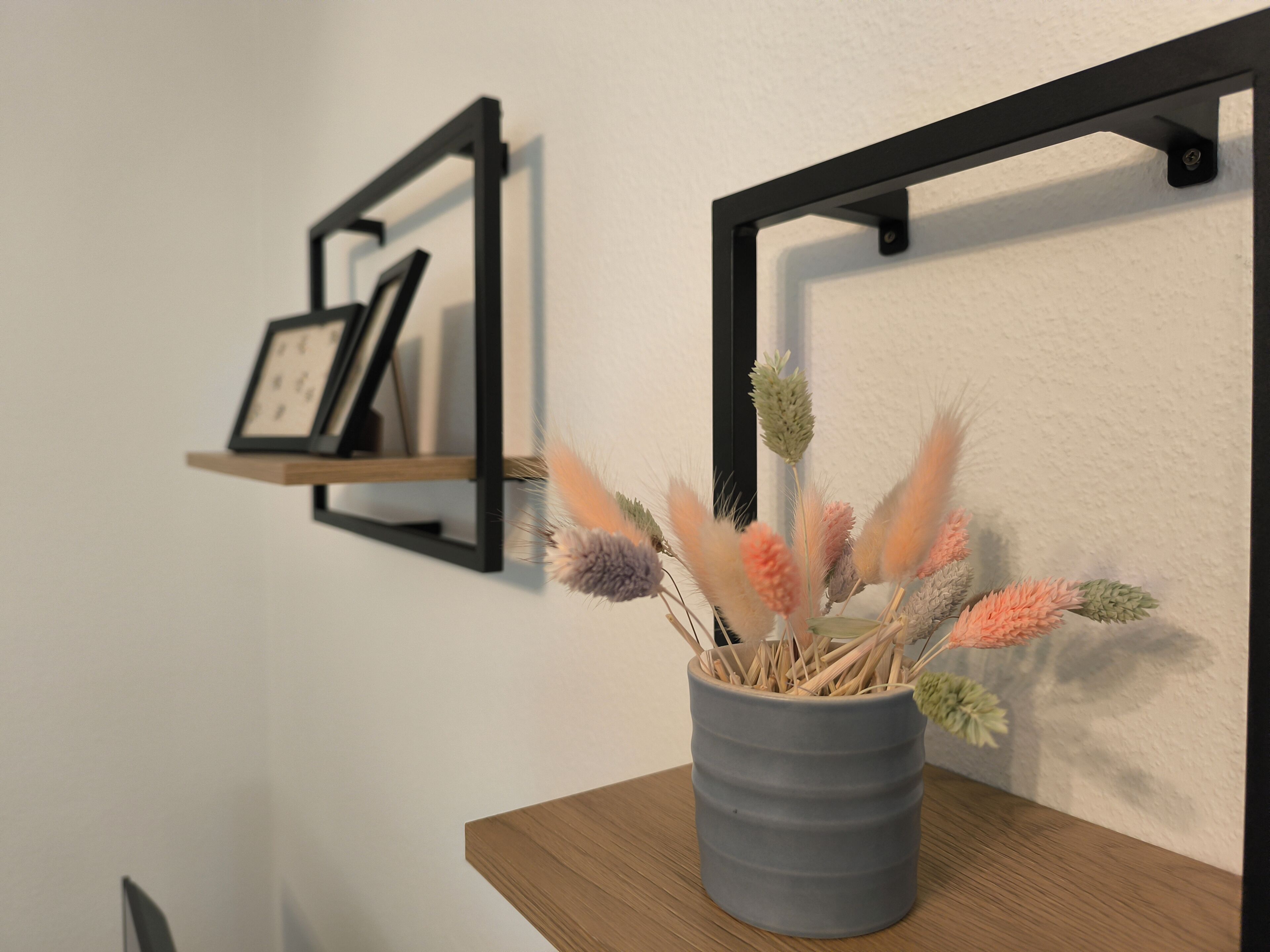 Een grijze vaas met gedroogde bloemen op een houten tafel tegen een witte muur.