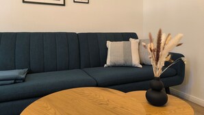 A modern living room with a dark green sofa, two pillows, a round wooden coffee table, and a vase with dried plants.