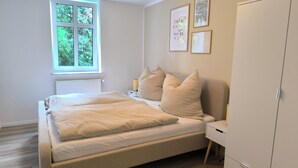 A bedroom with a bed, a nightstand, a wardrobe, a window with a view of greenery, and framed botanical prints on the wall.