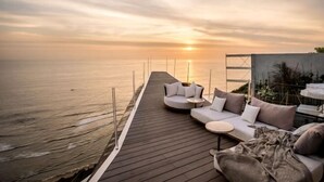 A wooden deck with a sofa and table, overlooking the sea at sunset. - Private Jet Villa Uluwatu (Bali)
