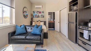 Eine moderne Wohnung mit einem Wohnzimmer, ausgestattet mit einem schwarzen Ledersofa, einem Couchtisch und einem großen Fenster. Die Küche beinhaltet einen Edelstahl-Kühlschrank und ein Kochfeld.