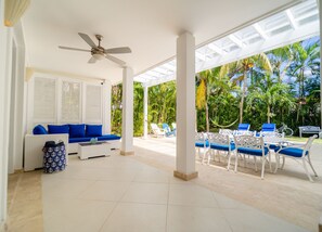 A covered outdoor seating area with a ceiling fan, a sofa with blue cushions, and a table with chairs.